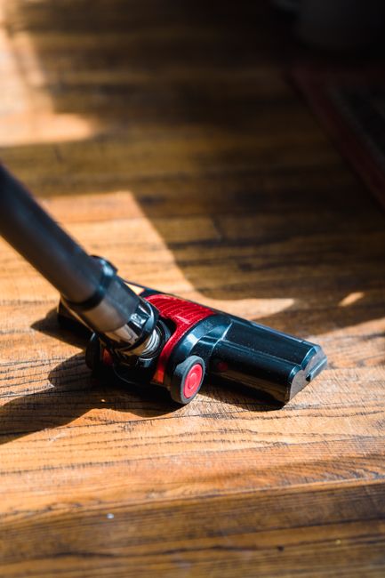 A close-up view of a handheld vacuum cleaner with a black body and red accents, positioned on a wooden outdoor deck surface. The vacuum's nozzle is resting on the textured, weathered wood planks, whic